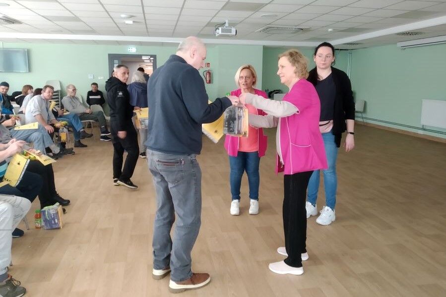 Grupa kobiet i mężczyzn stojących w pomieszczeniu. Na pierwszym planie kobieta wręcza mężczyźnie prezent i dyplom.