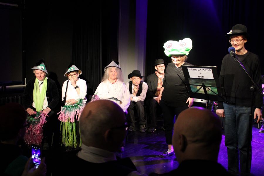 Grupa pięciu kobiet w kostiumach teatralnych i wyszukanych nakryciach głowy. W tle dwaj siedzący mężczyźni.