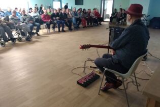 Grupa kobiet i mężczyzn siedzących na krzesłach w pomieszczeniu. Na pierwszym planie mężczyzna z gitarą przy mikrofonie.