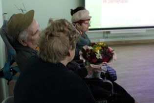 Sfotografowana tyłem siedząca kobieta z bukietem w dłoniach. Na drugim planie siedzący kobieta i mężczyzna.