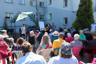 Grupa kobiet i mężczyzn będących publicznością w plenerze na tle budynku. Pośrodku dwaj mężczyźni przy mikrofonie, z których jeden gra na gitarze.