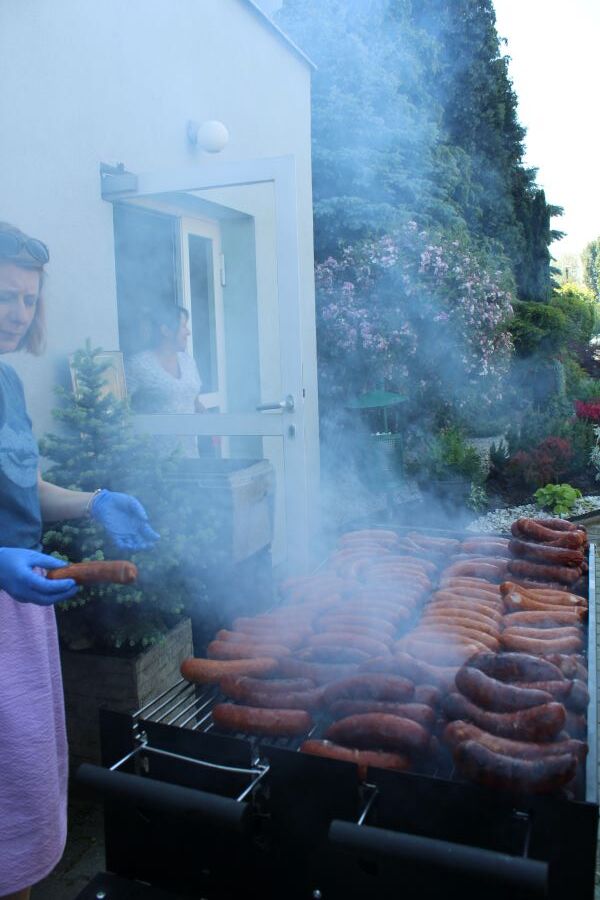Grill z kiełbaskami przed budynkiem na tle zieleni. Nad potrawą unoszą się kłęby dymu.