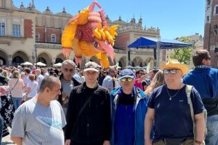 Grupa kobiet i mężczyzn w plenerze na tle zabytkowych budowli. Na pierwszym planie pięciu stojących mężczyzn, a nad nimi unoszący się w powietrzu smok