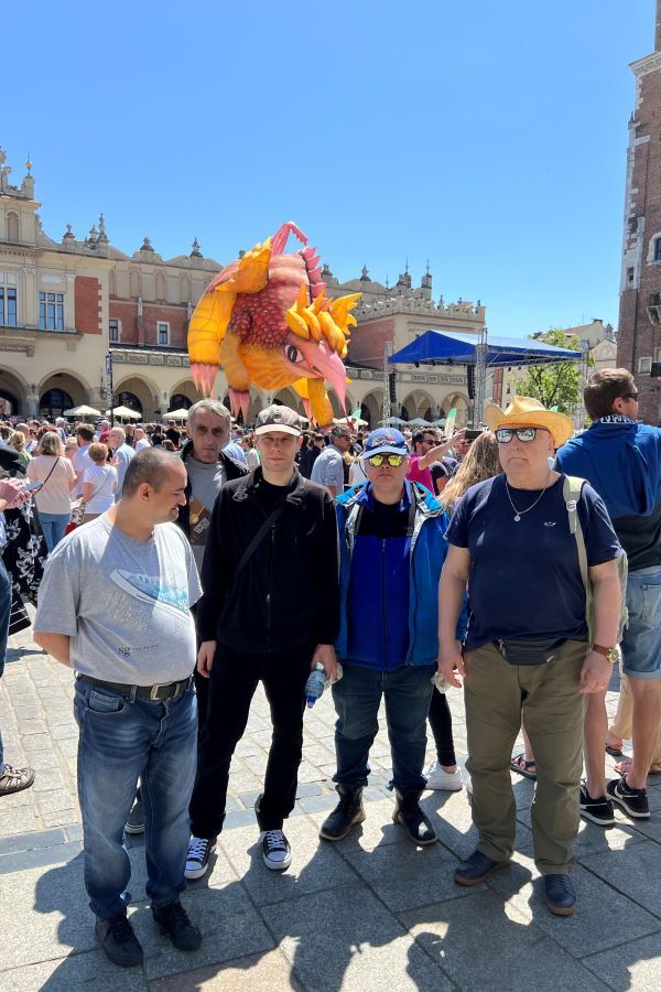 Grupa kobiet i mężczyzn w plenerze na tle zabytkowych budowli. Na pierwszym planie pięciu stojących mężczyzn, a nad nimi unoszący się w powietrzu smok
