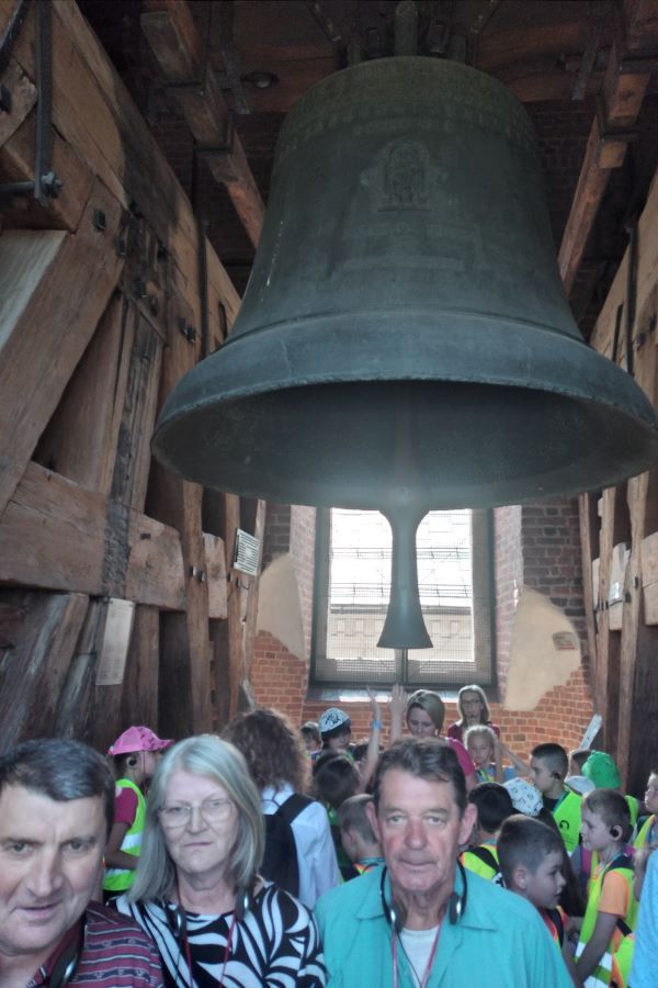 Grupa kobiet i mężczyzn oraz dzieci w pomieszczeniu. W centrum nad ich głowami wisi dzwon.