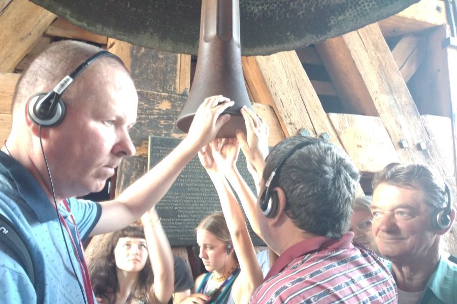 Grupa kobiet i mężczyzn w słuchawkach na tle drewnianej konstrukcji. Wszyscy dotykają wyciągniętymi w górę dłońmi serca dzwonu.