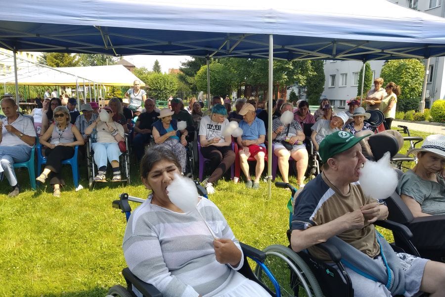 Grupa siedzących kobiet i mężczyzn w plenerze. Na pierwszym planie kobieta i mężczyzna jedzą watę cukrową.