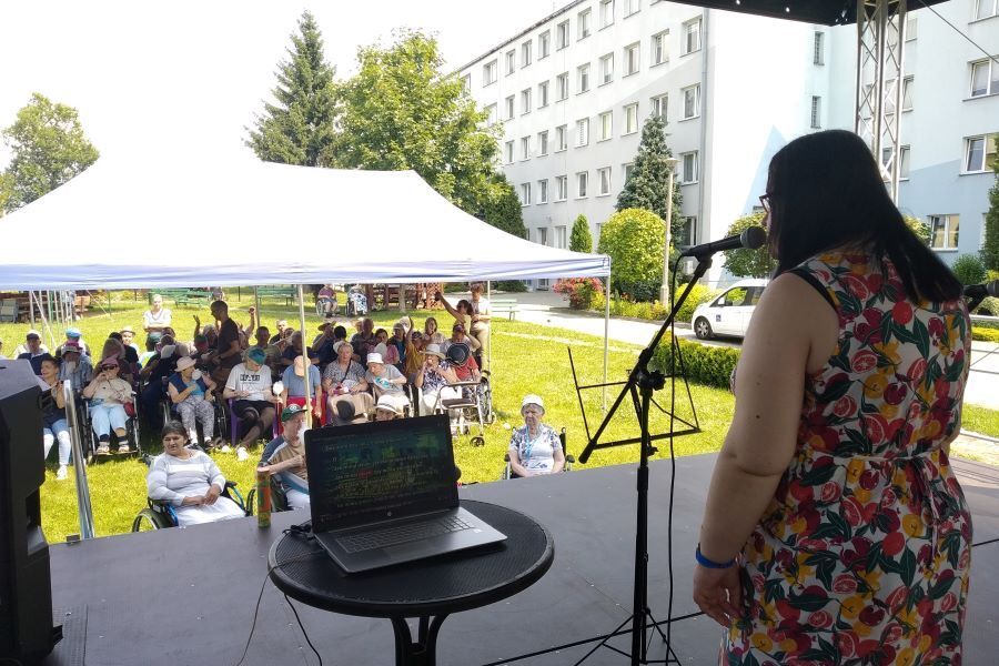 Kobieta przy mikrofonie na scenie plenerowej. Przed nią grupa siedzących kobiet i mężczyzn w cieniu pawilonu ogrodowego.