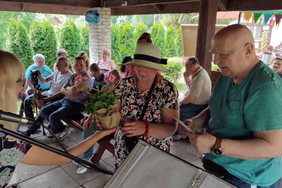 Grupa kobiet i mężczyzn siedzących w altanie. W centrum dwie kobiety i mężczyzna, jedna z pań wręcza drugiej pelargonię w donicy. 