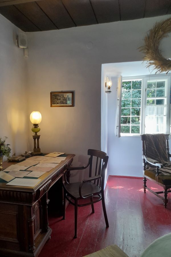 Zabytkowy gabinet we wnętrzach dworku. Biurko, na którym ustawiona jest zapalona lampa, dwa fotele i obraz na ścianie. W tle otwarte okno.