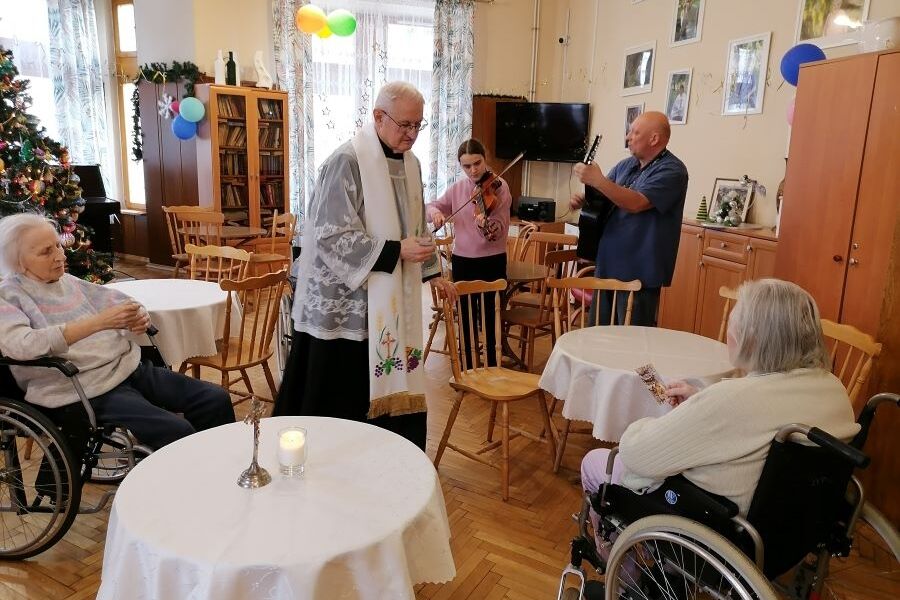 Ksiądz, mieszkanki Domu oraz pracownicy grający kolędy na instrumentach podczas Kolędy noworocznej w świetlicy Domu.