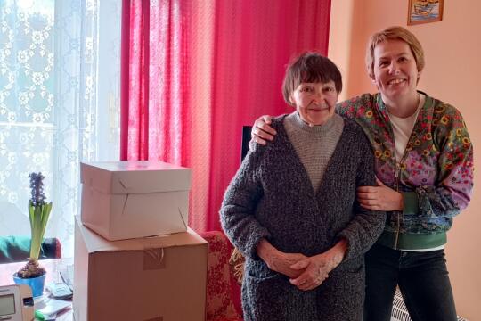 Starsza Pani w mieszkaniu obok pracownik socjalny widoczne pudła z żywnością