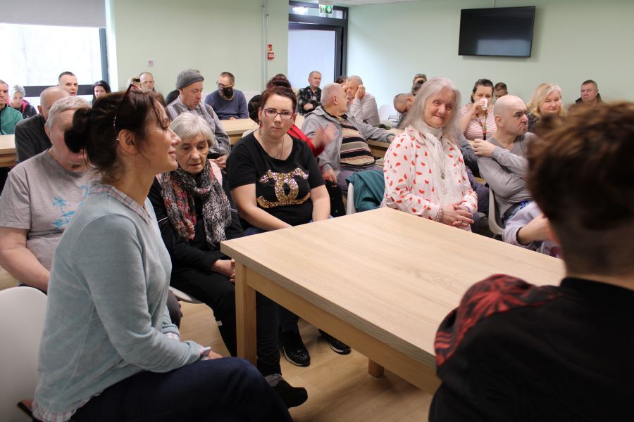 Grupa kobiet i mężczyzn siedzących przy stołach w pomieszczeniu.