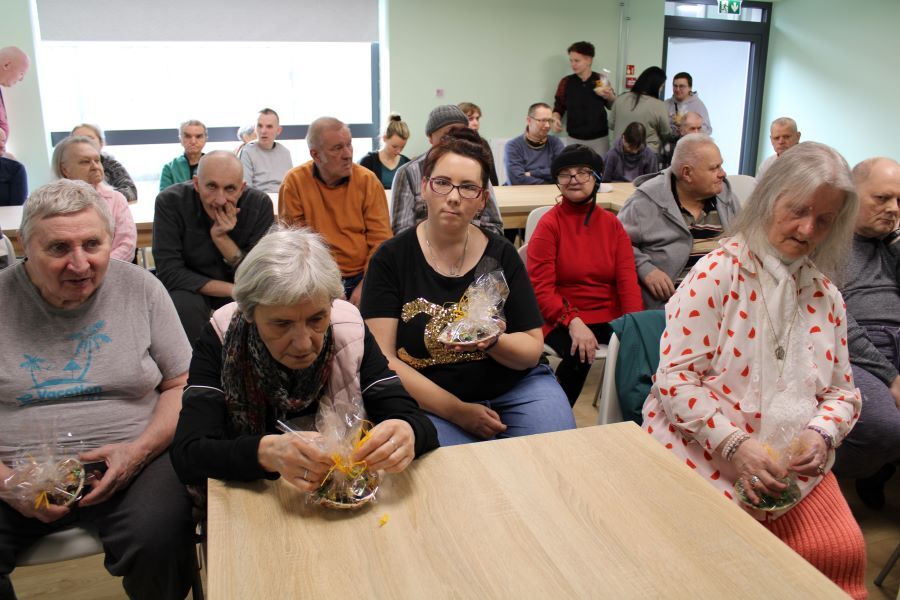Trzy kobiety i mężczyzna rozpakowują prezenty. W tle grupa kobiet i mężczyzn siedzących przy stole.