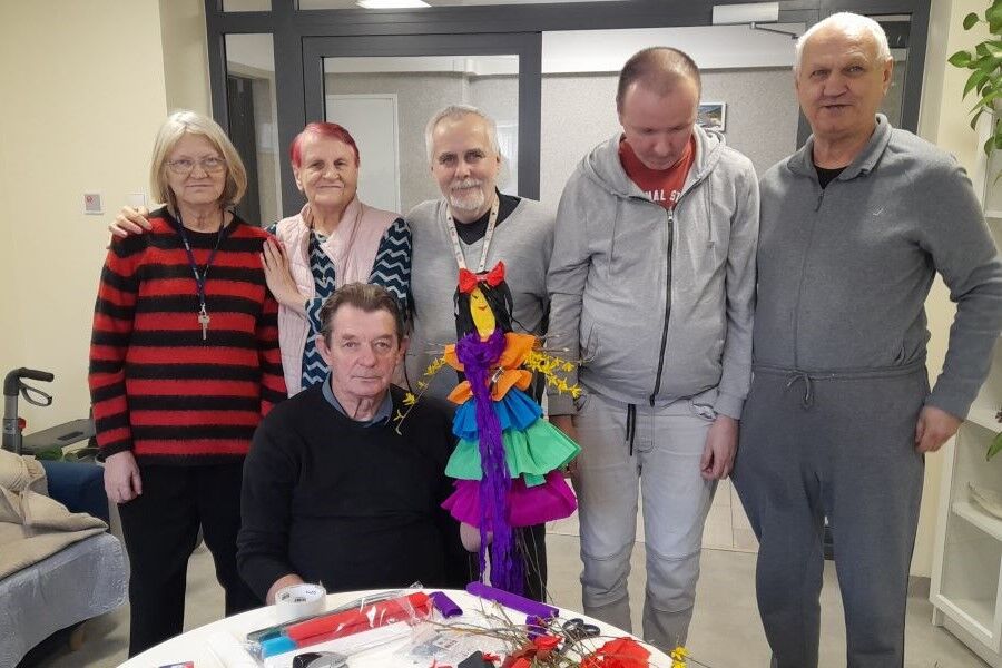 Dwie kobiety i czterech mężczyzn w pomieszczeniu. Jeden z panów siedzi przy stole i trzyma w dłoni kukłę z bibuły.