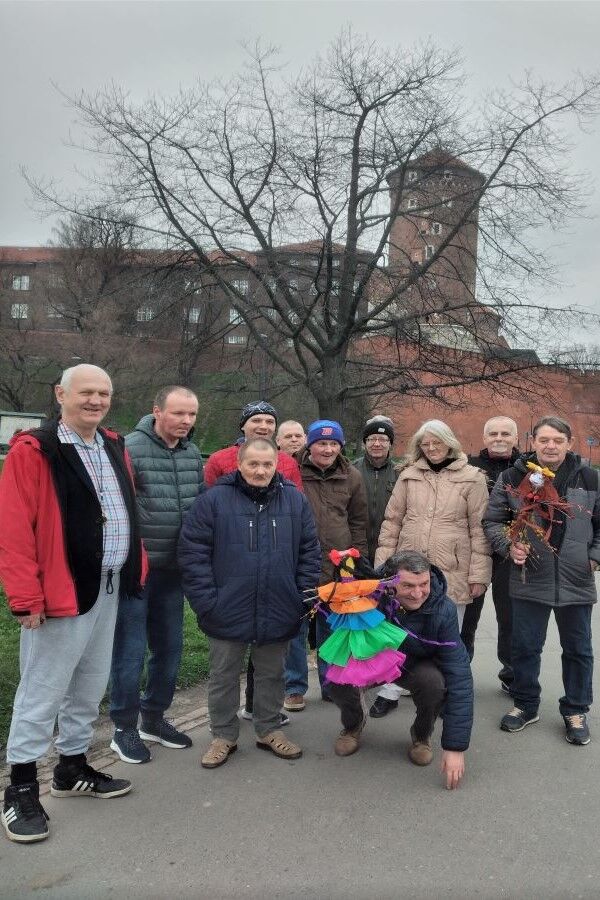 Kobieta i dziesięciu mężczyzn stojących na tle zabudowań zamku. Dwaj panowie trzymają w rękach kukły z patyków i bibuły.