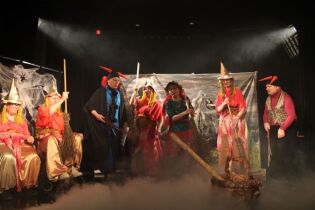 Pan Twardowski Grupa kobiet i mężczyzn w kostiumach teatralnych na oświetlonej scenie.