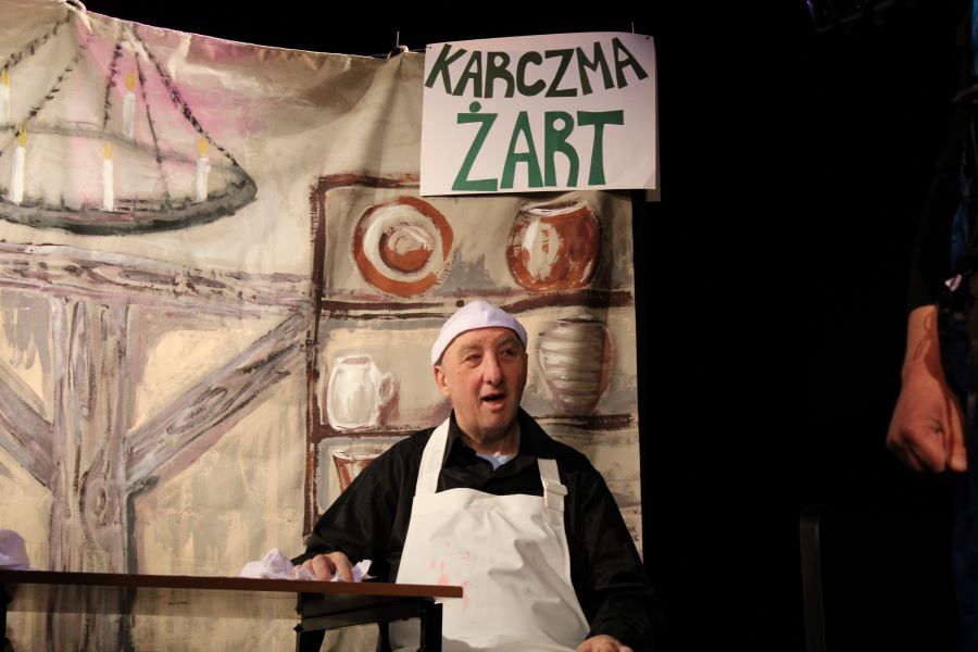 Mężczyzna siedzący przy stole na tle scenografii teatralnej.