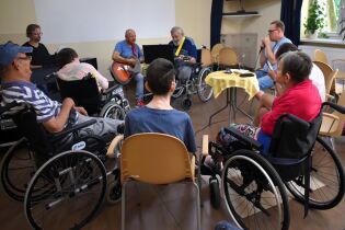 Kilku pracowników i mieszkaniec grają na instrumentach, inni mieszkańcy przysłuchują się.