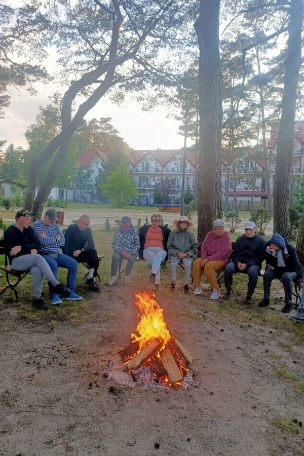 Pięć kobiet i czterech mężczyzn siedzących na ławkach przy ognisku. W tle drzewa i budynki.