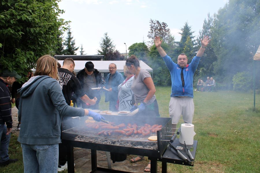 Grupa kobiet i mężczyzn w plenerze. Na pierwszym planie grill ze smażącymi się kiełbasami.