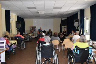 Zajęcia muzyczne 27.06.2024 Mieszkańcy siedzą na świetlicy domu, słuchają jak terapeuta wraz z jednym z mieszkańców gra na gitarze i śpiewa.