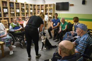 Zajęcia sensoryczne 04.07.2024 Kilkunastu mieszkańców siedzi w kręgu. W środku opiekunka wraz z psem rasy border collie.