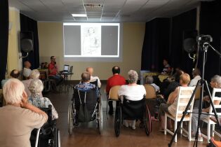 Spotkania historyczne 12.07.2024 Kilkunastu mieszkańców siedzi przed dużym ekranem, z boku Pan Dyrektor trzyma mikrofon.