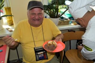 Zajęcia kulinarne 26.07.2024 Mieszkaniec siedzi na stołku, trzyma talerzyk z szarlotką i lodami, szeroko się uśmiecha.