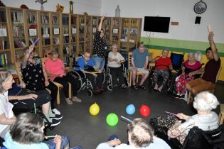 Zajęcia sensoryczne 01.08.2024 Kilkunastu mieszkańców siedzi w kręgu, podnoszą do góry świecące pałeczki.
