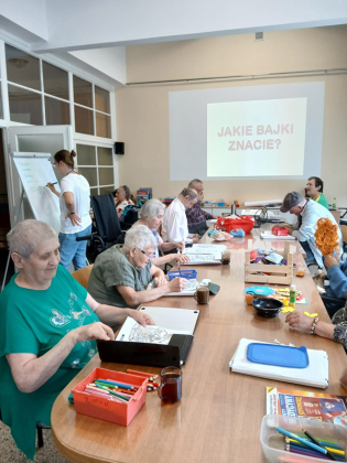 Terapia dopołudniowa 05.08.2024 Mieszkańcy siedzą zgromadzeni przy dużym stole. Na ścianie wyświetlany jest temat zajęć - Bajki