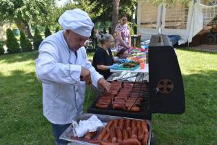 Grill 07.08.2024 Mężczyzna w stroju kucharza stoi przy grillu i smaży kiełbaski.