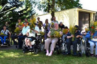 Bukiety 14.08.2024 Kilkunastu mieszkańców, Dyrekcja i pracownicy pozują w ogrodzie, trzymając bukiety z ziół.