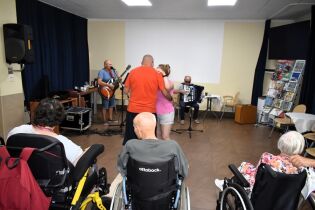 Zajęcia muzyczne 16.08.2024 Pracownik z wolontariuszem grają na gitarze i akordeonie, para mieszkańców tańczy a kilku mieszkańców obserwuje.