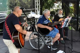 Piknik 21.08.2024 Mieszkaniec na wózku gra na gitarze na estradzie, towarzyszy mu dwóch terapeutów.