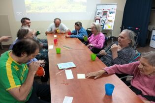 Neurobik 03.09.2024 Kilkunastu mieszkańców siedzi przy długim stole, dmuchają przez rurki.