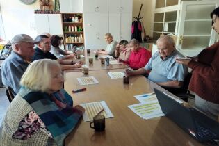 Zajęcia poznawcze 14.10.2024 Kilkunastu mieszkańców siedzi przy długim stole, pani psycholog czyta im zagadki .