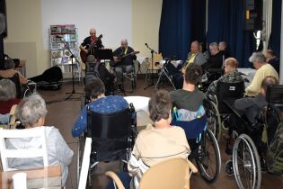 Zajęcia muzyczne 17.10.2024 Kilkunastu mieszkańców siedzi wokół terapeuty i mieszkańca grających na gitarach.