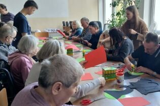 Arteterapia 22.11.2024 Kilkunastu mieszkańców siedzi przy długim stole, wycinają kolorowy papier.