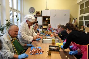 Zajęcia kulinarne 28.11.2024 Kilkunastu mieszkańców siedzi przy długim stole, kroją produkty na koreczki.