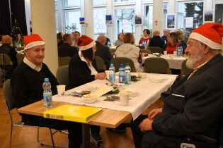 Świąteczne spotkanie 12.12.2024 Troje mieszkańców odświętnie ubranych siedzi przy stole, w tle duży stół i inni goście.