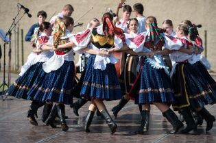 Grafika przedstawia chłopców i dziewczęta, ubranych w stroje regionalne, tańczących w kółku taniec na scenie.