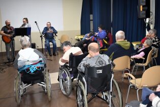 Muzykoterapia 13.02.2025 Dwoje terapeutów śpiewa, kilkunastu mieszkańców słucha piosenek.