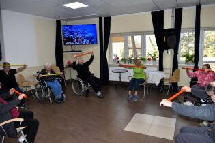 Gimnastyka 14.02.2025 Kilkoro mieszkańców siedzi w kręgu, trzymają kolorowe laski gimnastyczne .