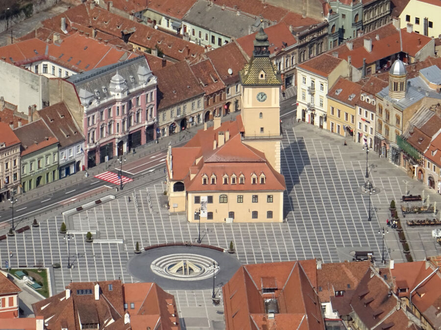 Zdjęcie przedstawia rynek w Braszowie z lotu ptaka. Widoczny jest rynek w kształcie trójkąta i okalające go kamienice i nowoczesna okrągła fontanna a na środku zabytkowy ratusz miejski z wieżą.