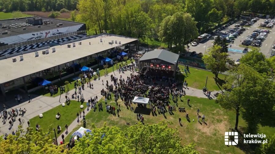 Widok z góry na plenerowe wydarzenie przy Nowohuckim Centrum Kultury w Krakowie: scena z koncertem, tłum ludzi, namioty wystawców, zielony teren z drzewami i parking w tle, słoneczna pogoda.
