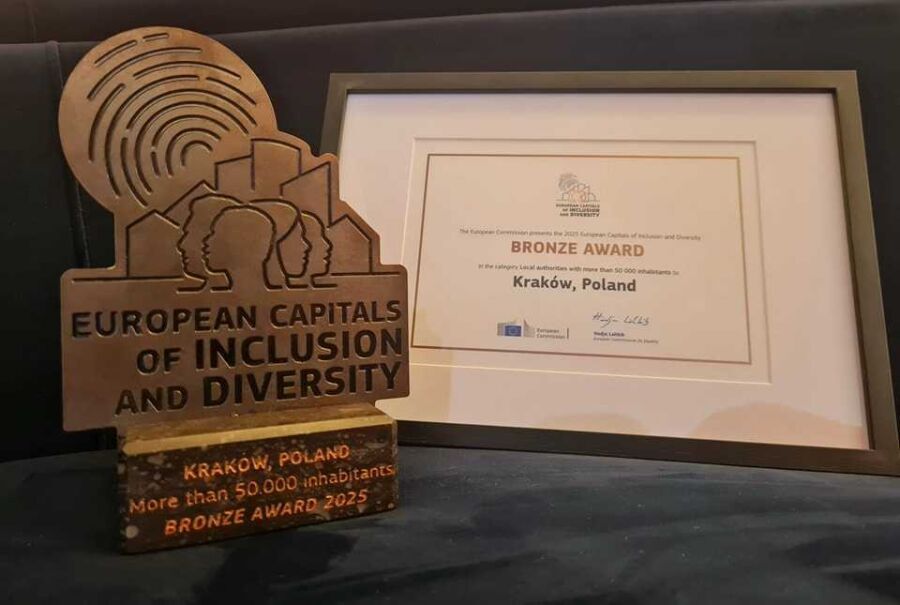 Kraków z nagrodą Komisji Europejskiej za działania na rzecz różnorodności i inkluzywności. Po lewej stronie metalowa statuetka nagrody z metalu w kolorze starego brązu a po prawej nieco w głębi kremowy dyplom dla Krakowa oprawiony w ciemnobrązową ramkę.