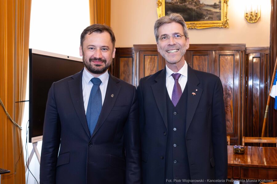 Wizyta Ambasadora Austrii u Prezydenta Krakowa 5 maja 2025 r. Na fotografii Prezydent Aleksander Miszalski po lewej oraz Ambasador Austrii po prawej pozują do fotografii ustawieni przodem  na tle ciemnej brązowej boazerii i flag miasta w gabinecie Prezydenta Krakowa. 