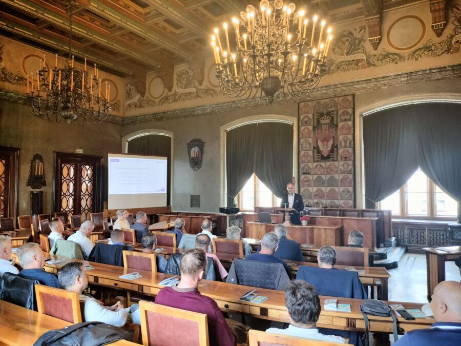 Na zdjęciu widoczna od tyłu grupa mężczyzn zasiadająca w sali Obrad Rady Miasta Krakowa. W głębi w centralnej części zdjęcia widoczna mównica z której przemawia Sekretarz Miasta Antoni Fryczek. Po lewej stronie w głębi widoczny ekran na którym wyświetlany jest slajd prezentacji z tekstem.