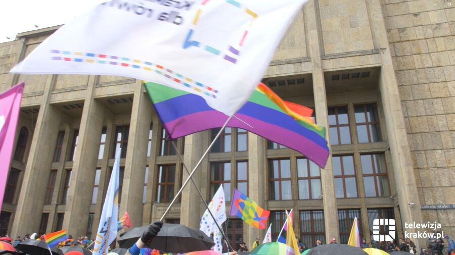 Tłum ludzi z parasolkami i tęczowymi flagami przed Muzeu Narodowego w Krakowie - Gmach Główny. Widoczne różnorodne flagi społeczności LGBTQ+. W prawym dolnym rogu logo „telewizja kraków.pl”. Zdjęcie zrobiono podczas Marszu Równości.